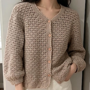 Può includere: Cardigan beige all'uncinetto con trama aperta, scollo a V e maniche lunghe. Il cardigan ha una chiusura con bottoni con quattro bottoni rotondi marrone chiaro. Il motivo all'uncinetto è un disegno geometrico ripetuto. Le maniche sono leggermente a sbuffo sulle spalle.