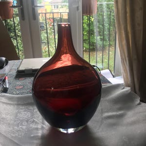 Può includere: Un vaso in vetro rosso scuro traslucido con un corpo rotondo e bulboso e un collo stretto e allungato. Il vetro sfuma da un cremisi scuro e saturo alla base verso una tonalità di rosso più chiara e trasparente vicino all'apertura superiore.