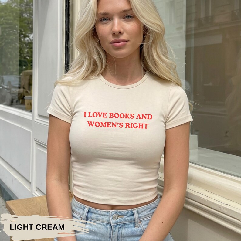 Może przedstawiać: Jasnokremowa koszulka z kr&oacute;tkim rękawem z czerwonym napisem "I LOVE BOOKS AND WOMEN'S RIGHT". Koszulka ma dopasowany kr&oacute;j i jest połączona z niebieskimi dżinsami. Na dole obrazu widnieje napis "LIGHT CREAM".
