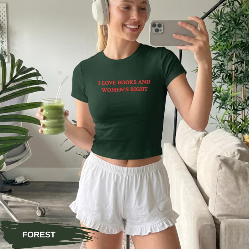 Może przedstawiać: Leśnozielony top z napisem "I LOVE BOOKS AND WOMEN'S RIGHT" w kolorze czerwonym. Modelka ma na sobie białe szorty. Na zdjęciu znajduje się r&oacute;wnież zielony nap&oacute;j w szklance i białe słuchawki.