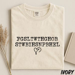 May include: An ivory short-sleeved t-shirt featuring black text that reads FGSLTWTHGHOB STWBIHSNPBHEL with a heart and cross symbol underneath. The shirt has a crew neckline and a Comfort Colors brand label printed on the inside of the collar.