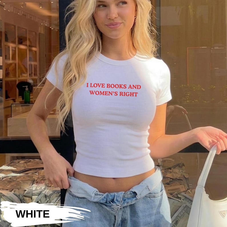 Może przedstawiać: Biały t-shirt z kr&oacute;tkim rękawem z czerwonym napisem "I LOVE BOOKS AND WOMEN'S RIGHT". Koszulka ma dopasowany kr&oacute;j i jest połączona z jasnymi dżinsami. Osoba trzyma białą torebkę.