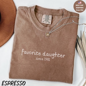 May include: Espresso-coloured t-shirt with the words "favorite daughter since 1981" in white script. A silver necklace with a pendant rests on the shirt. The shirt has a crew neck and a Comfort Colors tag.