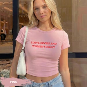 Może przedstawiać: R&oacute;żowa koszulka z kr&oacute;tkim rękawem z czerwonym napisem "I LOVE BOOKS AND WOMEN'S RIGHT". Koszulka ma kr&oacute;tki kr&oacute;j. Modelka ma na sobie niebieskie dżinsy i białą torebkę. Koszulka jest wykonana z miękkiego materiału.