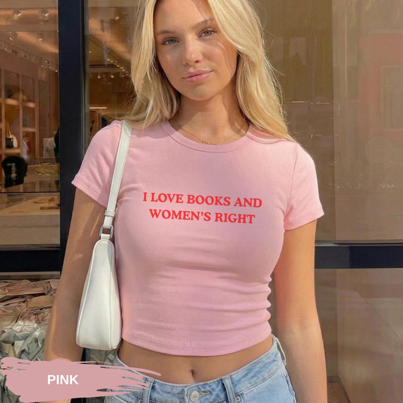 Może przedstawiać: R&oacute;żowa koszulka z kr&oacute;tkim rękawem z czerwonym napisem "I LOVE BOOKS AND WOMEN'S RIGHT". Koszulka ma kr&oacute;tki kr&oacute;j. Modelka ma na sobie niebieskie dżinsy i białą torebkę. Koszulka jest wykonana z miękkiego materiału.