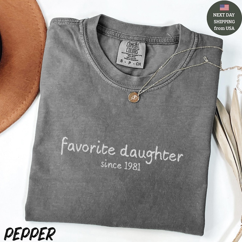 May include: A grey Comfort Colors t-shirt with the text "favorite daughter since 1981" in white script. A silver necklace with a pendant rests on the shirt. The shirt is folded, and the word "PEPPER" is at the bottom.