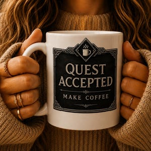 Könnte beinhalten: Ein weißer Keramikbecher mit einer schwarzen rechteckigen Grafik und einem dekorativen Rand. Der Text lautet QUEST ACCEPTED in großen Buchstaben und MAKE COFFEE in kleineren Buchstaben darunter, mit einem Symbol einer dampfenden Tasse.