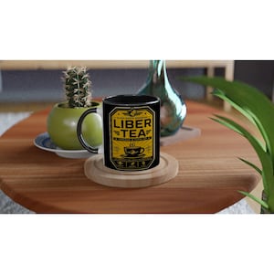 Könnte beinhalten: Eine schwarze Keramiktasse mit einem gelb-schwarzen Etikett mit der Aufschrift "Liber Tea" und einer Teetassengrafik. Die Tasse hat einen schwarzen Henkel und Rand. Die Tasse steht auf einem Holzuntersetzer.