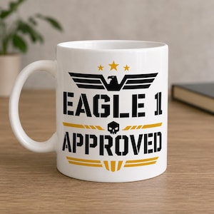 Könnte beinhalten: Weiße Keramiktasse mit dem Text "EAGLE 1 APPROVED" in Schwarz und Gelb. Das Design umfasst ein Adleremblem, Sterne und einen Totenkopf. Die Tasse hat einen gebogenen Henkel und steht auf einer Holzoberfläche.