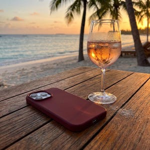 May include: A glass of rose wine with ice sits on a wooden table next to a maroon phone case. The background features a beach, ocean, and palm trees at sunset. The scene evokes relaxation and leisure.