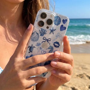 Peut inclure: Coque de téléphone transparente avec un motif nautique bleu et blanc. Le design comprend des illustrations de coquillages, d'étoiles de mer, d'hippocampes, de nœuds, de cœurs et d'une tortue. En arrière-plan, une plage et la mer.