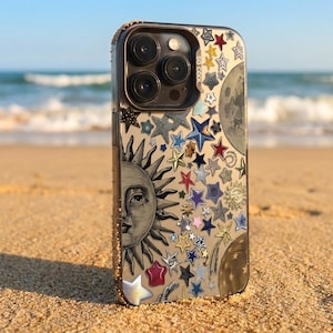 Op de afbeelding: Transparante telefoonhoes versierd met verschillende stervormen, een zon en een maan. De hoes heeft een gouden glitterrand en de tekst "SO COOL" is zichtbaar. De hoes ligt op een zandstrand met de oceaan op de achtergrond.