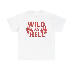 Pode incluir: Uma t-shirt branca de manga curta com o texto vermelho WILD AS HELL impresso na frente. A palavra WILD está posicionada acima da palavra AS, e a palavra HELL está abaixo, com gráficos de chamas vermelhas estilizadas em ambos os lados.