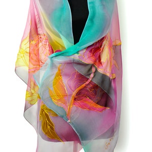 May include: A sheer silk scarf with a floral design in shades of pink, yellow, and turquoise. The scarf is hand-painted with gold accents and features a delicate floral pattern.