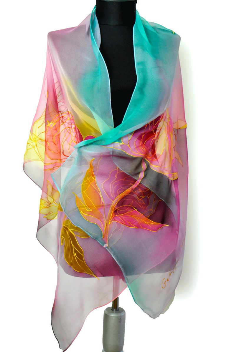 May include: A sheer silk scarf with a floral design in shades of pink, yellow, and turquoise. The scarf is hand-painted with gold accents and features a delicate floral pattern.