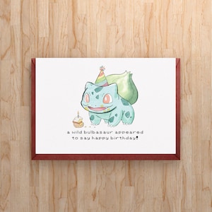 May include: A white greeting card with a red border featuring a watercolour illustration of a light blue Bulbasaur wearing a rainbow party hat next to a small cupcake with a candle. The text reads: a wild bulbasaur appeared to say happy birthday!