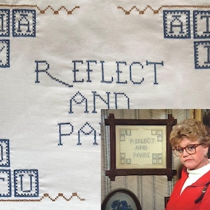 May include: A white cross-stitch sampler with the words "REFLECT AND PAUSE" in blue thread. The sampler has a brown border and blue letters in the corners. A framed sampler with the same text is in the image.