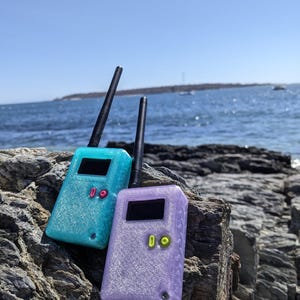 May include: Two glittery handheld radios, one teal and one lavender, rest on a rocky surface with the ocean in the background. Each radio has a black antenna, a small screen, and a button. The radios are a unique design.