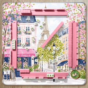Op de afbeelding: Een vierkante mahjongmat met een Parijse straatscène met de Eiffeltoren, roze kersenbloesems en een café. Vier roze rekjes zijn in een vierkant geplaatst, met verspreide witte mahjongstenen en een kleine ronde groene speelschijf.