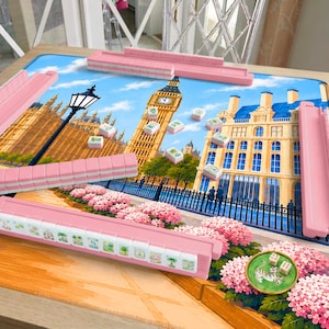 Peut inclure: Un jeu de mah-jong avec quatre supports de tuiles roses et des tuiles blanches aux motifs verts, disposé sur un tapis de jeu illustrant Londres avec Big Ben, un lampadaire et des hortensias roses, et un petit plat rond vert avec deux dés.