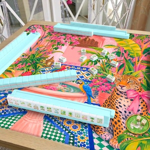 Könnte beinhalten: Ein Mahjong-Set mit vier hellblauen Fliesenhaltern und einer bunten Spielmatte mit tropischem Dschungel-Druck. Die Matte zeigt einen Leoparden mit rosa Schleife, einen blau-gelben Ara, einen Brunnen und grünes Blattwerk. Verstreute Mahjong-Steine und zwei Würfel liegen bereit.
