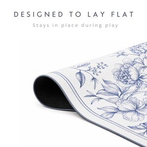 May include: A white mat with a blue floral pattern and a dark underside, featuring the text "DESIGNED TO LAY FLAT" and "Stays in place during play" printed above the mat. The mat is shown partially rolled to reveal the dark, textured backing.