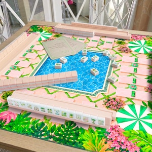May include: A Mahjong game set featuring pastel pink tile racks and tiles with botanical illustrations. The game board displays a vibrant swimming pool graphic surrounded by tropical foliage, green and white umbrellas, and pink flowers.