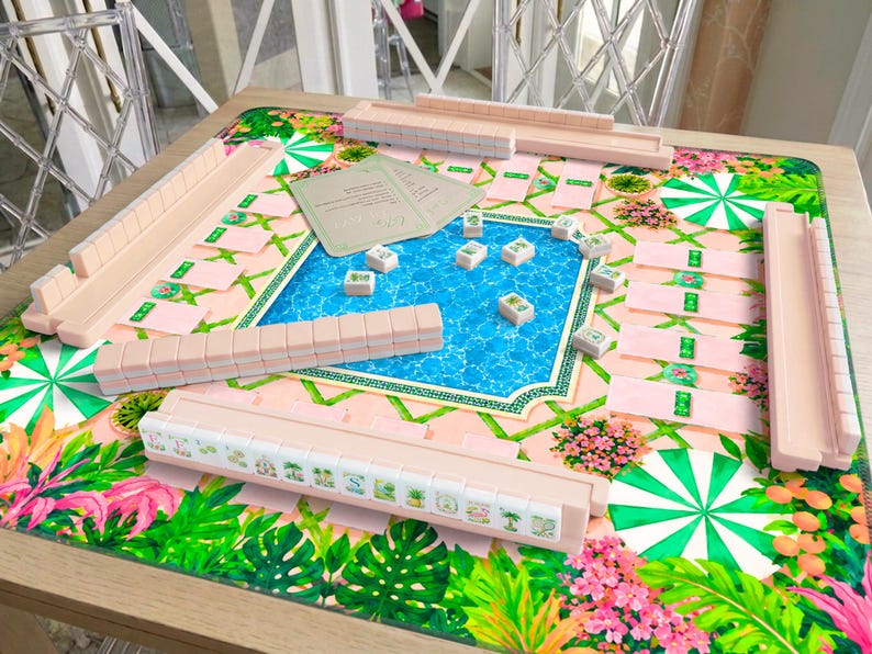 May include: A Mahjong game set featuring pastel pink tile racks and tiles with botanical illustrations. The game board displays a vibrant swimming pool graphic surrounded by tropical foliage, green and white umbrellas, and pink flowers.