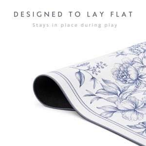 May include: A white mat with a blue floral pattern and a dark underside, featuring the text "DESIGNED TO LAY FLAT" and "Stays in place during play" printed above the mat. The mat is shown partially rolled to reveal the dark, textured backing.