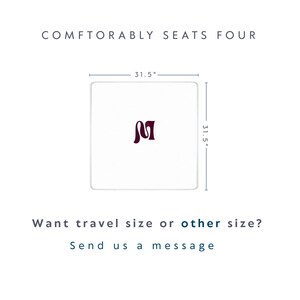 May include: A square white table with a dark maroon letter M in the center. The table measures 31.5 inches by 31.5 inches. Text above reads COMFTORABLY SEATS FOUR. Text below reads Want travel size or other size? Send us a message.