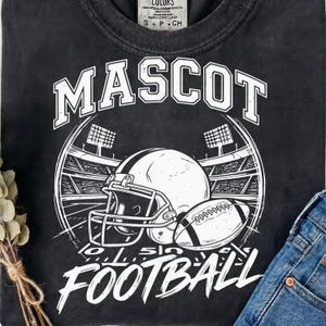 Puede incluir: Una camiseta negra con un estampado gráfico blanco que muestra un casco de fútbol americano y un balón frente a un estadio. El texto MASCOT aparece arqueado sobre el casco y la palabra FOOTBALL está impresa en letra grande en la parte inferior.