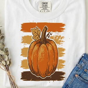 Peut inclure: Un t-shirt blanc avec un motif représentant une grosse citrouille orange avec une feuille et une tige. La citrouille est centrée sur cinq coups de pinceau horizontaux au style vieilli dans des tons orange, doré et marron. L'étiquette indique Comfort Colors.