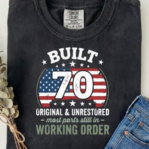 Peut inclure: Un t-shirt noir à manches courtes avec un motif graphique portant le texte "BUILT 70 ORIGINAL & UNRESTORED most parts still in WORKING ORDER" sur un drapeau américain stylisé. Le t-shirt présente un col rond et une étiquette Comfort Colors.