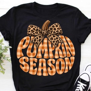 Peut inclure: Un t-shirt noir à manches courtes orné d'un grand motif de citrouille à carreaux vichy marron et blanc. La citrouille est surmontée d'un nœud à imprimé léopard et porte l'inscription PUMPKIN SEASON dans une police grasse et arrondie.
