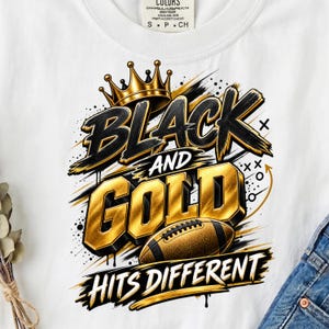 May include: A white t-shirt featuring a graphic design with a gold crown, a brown football, and the text "BLACK AND GOLD HITS DIFFERENT" in bold black and gold lettering, accented with black paint splatters and abstract line markings.