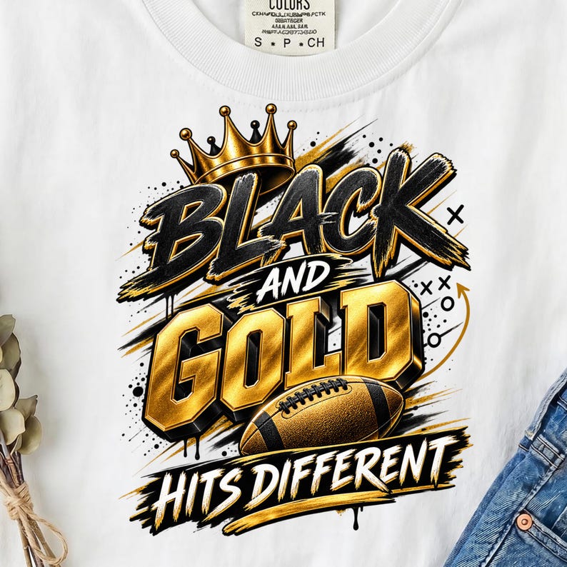 May include: A white t-shirt featuring a graphic design with a gold crown, a brown football, and the text "BLACK AND GOLD HITS DIFFERENT" in bold black and gold lettering, accented with black paint splatters and abstract line markings.