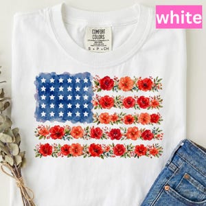 Peut inclure: T-shirt blanc avec un motif de drapeau américain floral. Les étoiles du drapeau sont blanches, les rayures sont composées de fleurs rouges et orange, et le fond est bleu aquarelle. Le t-shirt a un col rond et des manches courtes.