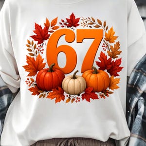 Peut inclure: Un t-shirt blanc orné d'un graphique représentant le nombre 67 en grande police orange, entouré d'une couronne circulaire de feuilles d'érable automnales colorées, de petites baies rouges et de trois citrouilles orange et crème.