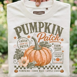 Peut inclure: Un t-shirt blanc cassé avec un motif représentant une grosse citrouille orange accompagnée de feuilles et de fleurs vertes. Le texte indique PUMPKIN Patch, FRESH FROM THE PATCH, FARM FRESH, LOCALLY GROWN, AUTUMN PICKED, PUMPKINS, CORN MAZE et APPLE CIDER.