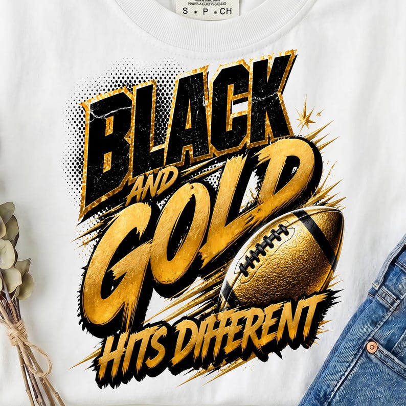 May include: A white t-shirt featuring a graphic design with the text "BLACK AND GOLD HITS DIFFERENT" in bold black and gold lettering. The design includes a textured gold football illustration with black laces, set against a dynamic background.
