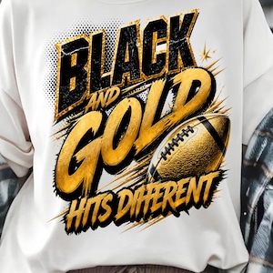May include: A white t-shirt featuring a graphic design with the text "BLACK AND GOLD HITS DIFFERENT" in bold black and gold lettering. The design includes a textured gold American football illustration with black laces and dynamic brush strokes.