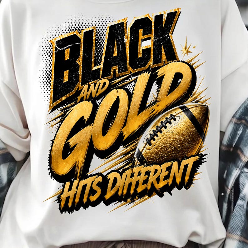 May include: A white t-shirt featuring a graphic design with the text "BLACK AND GOLD HITS DIFFERENT" in bold black and gold lettering. The design includes a textured gold American football illustration with black laces and dynamic brush strokes.