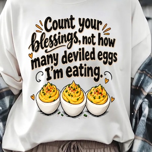 May include: A white sweatshirt featuring the black text "Count your blessings, not how many deviled eggs I'm eating." Below the text are three illustrations of deviled eggs with yellow yolks, green garnish, and orange heart accents.