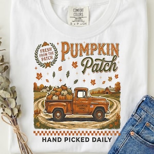 Peut inclure: Un t-shirt blanc avec un motif représentant un pick-up orange chargé de citrouilles sur un chemin de terre. Le texte indique Fresh From The Patch, Pumpkin Patch, Oakwood Farms et Hand Picked Daily, entouré de feuilles d'automne et d'une grange.