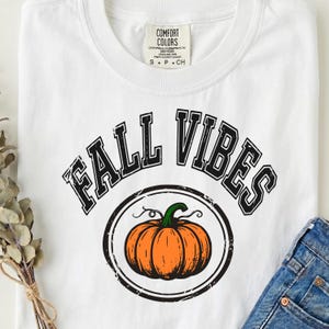 Peut inclure: Un t-shirt blanc à manches courtes avec le texte noir FALL VIBES en police arquée au-dessus d'un graphique circulaire contenant une citrouille orange avec une tige verte. Le t-shirt porte une étiquette de marque Comfort Colors à l'intérieur.