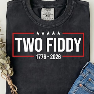 May include: A black crew neck t-shirt with a graphic print featuring a red rectangular border, five white stars, and the white text "TWO FIDDY" above the years "1776 - 2026". The shirt has a Comfort Colors brand label at the inner neckline.