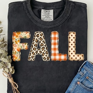Peut inclure: Un t-shirt noir à manches courtes avec un col rond portant le mot FALL imprimé en grandes lettres majuscules. Chaque lettre présente un motif distinct : floral, imprimé léopard, carreaux orange et blanc, et pois marron sur fond crème.