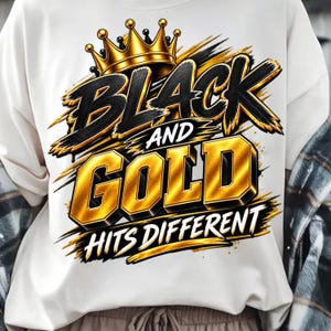 May include: A white sweatshirt featuring a graphic design with a gold crown above the text BLACK AND GOLD in bold, stylized lettering, followed by the text HITS DIFFERENT in a smaller font, all accented with black and gold brushstroke details.