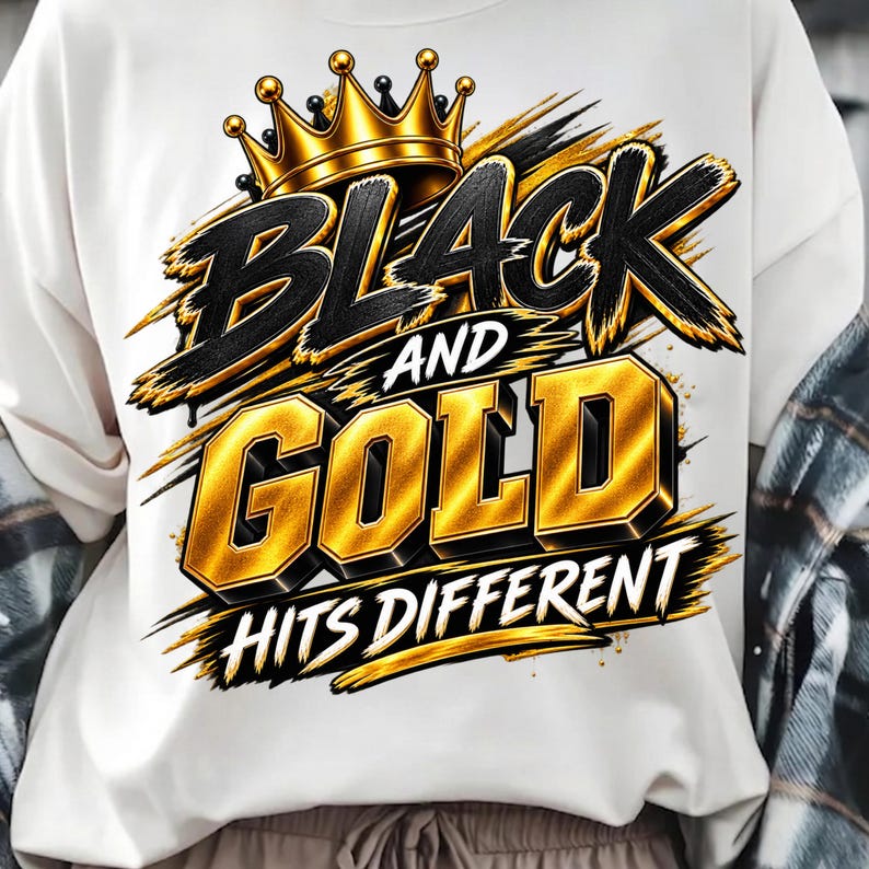 May include: A white sweatshirt featuring a graphic design with a gold crown above the text BLACK AND GOLD in bold, stylized lettering, followed by the text HITS DIFFERENT in a smaller font, all accented with black and gold brushstroke details.