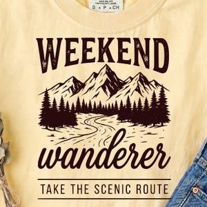 May include: A pale yellow t-shirt featuring a dark brown graphic of mountain peaks, a forest, and a winding path. The text reads WEEKEND wanderer and TAKE THE SCENIC ROUTE. The shirt has a crew neckline and a tag at the collar labeled S P CH.